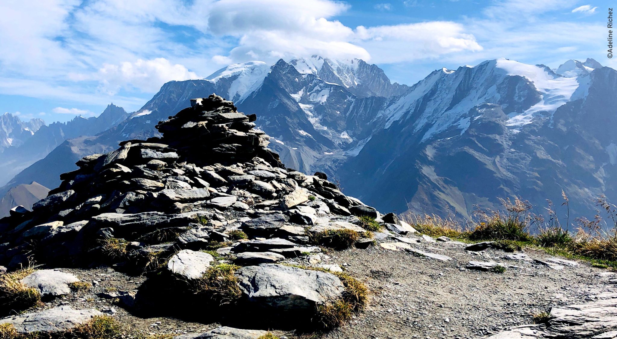 Au premier plan on voit un amas de pierre, un cairn, qui indique le sommet. Au second plan, on voit la chaine des sommets du massif du Mont Blanc.