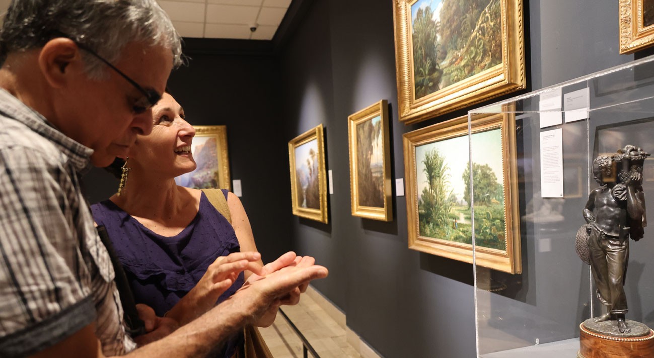 Deux personnes sont devant des tableaux au sein d'un musée.L'une d'elle dessine avec son doigt dans la main de l'autre.